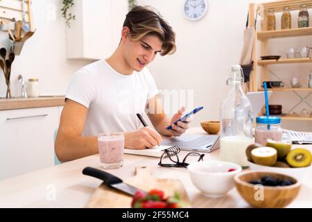 Young man with smart phone writing on notepad while sitting at table in kitchen Stock Photo