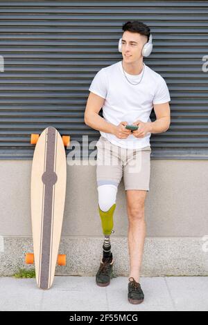 Young male amputee listening music while leaning on wall by skateboard Stock Photo