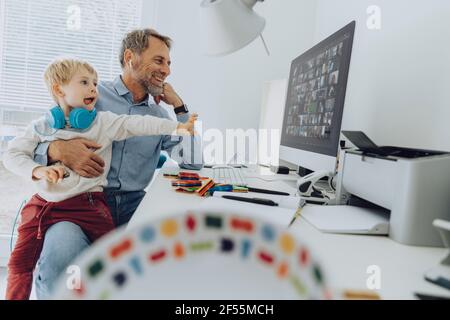 Playful son with wireless headphones sitting on lap of his father working at home office Stock Photo