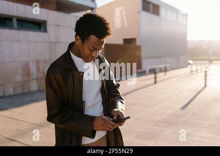 Mid adult man text messaging through smart phone while standing on footpath during sunset Stock Photo