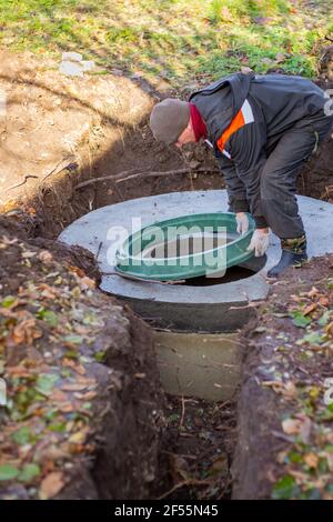 A worker installs a sewer manhole on a septic tank made of concrete rings. Construction of sewage disposal systems for private houses. Stock Photo