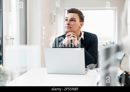Thoughtful businessman with laptop looking away while sitting at desk Stock Photo