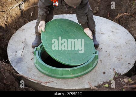 A worker installs a sewer manhole on a septic tank made of concrete rings. Construction of sewerage networks for country houses. Stock Photo