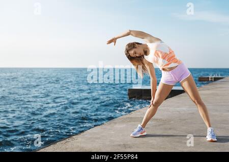 Sporty fit woman focused morning training session, doing runner stretch exercises, standing quay near sea raising hand, performing warm-up cardio Stock Photo