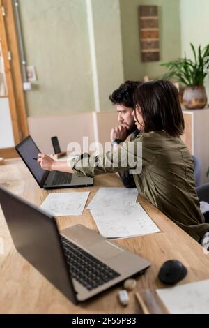 Female design professional assisting colleague while discussing over laptop at desk in industry Stock Photo