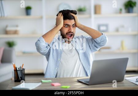 Deadline stress. Shocked arab man looking at laptop screen and touching head, suffering business problems Stock Photo