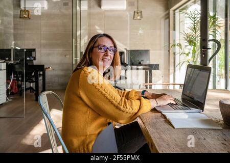 Mature female entrepreneur using laptop while sitting at conference table against glass door in office Stock Photo