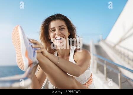 Happy healthy charming active fitness woman smiling, laughing joyfully stretching leg leaning quay bar, touching foot, warm-up before long distance Stock Photo