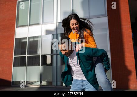 Man giving piggyback to cheerful girlfriend against modern building Stock Photo