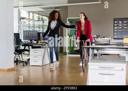 Cheerful female business professional assisting colleague while riding bicycle amidst desk in open plan office Stock Photo