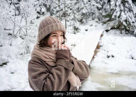 Happy woman wearing knit hat and jacket looking away while standing in forest Stock Photo