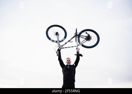 Cheerful sportsman lifting bicycle against sky Stock Photo