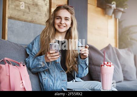 Good-looking fair-haired girl in trendy denim jacket sitting and relaxing in cafe, holding smartphone and credit card, wanting to pay for delicious Stock Photo