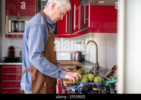 Senior man in apron cutting artichokes while doing video call with daughter on digital tablet at home Stock Photo