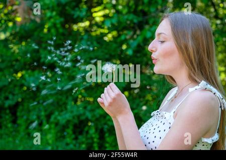 Blond teenage girl blowing dandelion while standing against plant Stock Photo