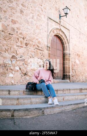 Smiling woman holding camera while sitting on staircase against wall Stock Photo