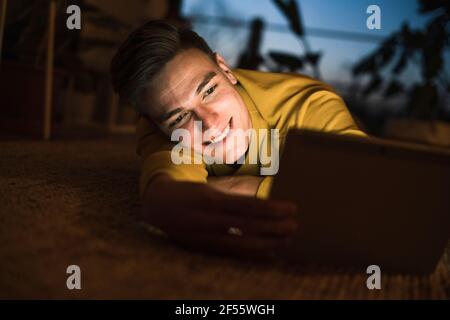 Smiling man using tablet while lying on carpet in living room during night Stock Photo