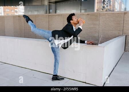 Flexible businessman with laptop drinking coffee while stretching leg Stock Photo