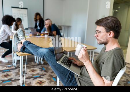 Male entrepreneur holding mug while working on laptop during meeting in office Stock Photo