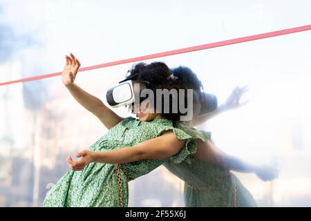 Young woman enjoying games wearing Virtual reality headset while leaning against glass wall Stock Photo