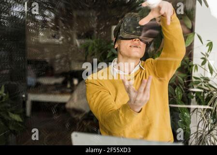 Young businessman gesturing while wearing virtual reality simulator seen through window in home office Stock Photo