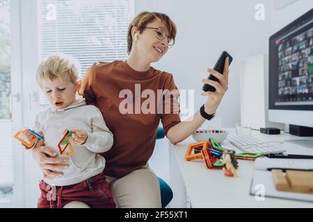 Mother using smart phone while son sitting on her lap playing with toys Stock Photo