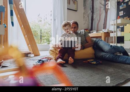Father lying on bean bag while looking at son using tablet in bedroom Stock Photo