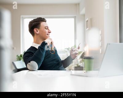 Smiling male entrepreneur surfing net through smart phone while sitting at home office Stock Photo