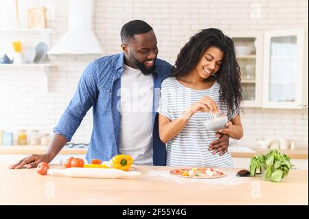 Happy african american couple cooking dinner together, trying new recipe where the main ingredient is love, woman adding grated cheese on dough for making delicious Italian pizza, husband supports her Stock Photo