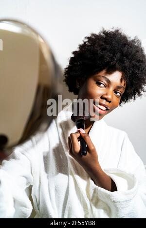 Young woman with hand mirror doing make-up while standing against white background Stock Photo