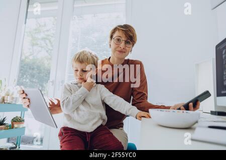 Smiling mother with son sitting on her lap while watching video on tablet at home office Stock Photo