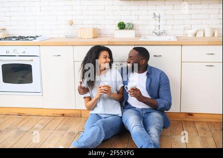 Young multiracial couple drinking coffee sitting on the kitchen floor, celebrate moving to a new apartment or buying new furniture with a cup of coffee tea, lovely girl in jeans talking with boyfriend Stock Photo