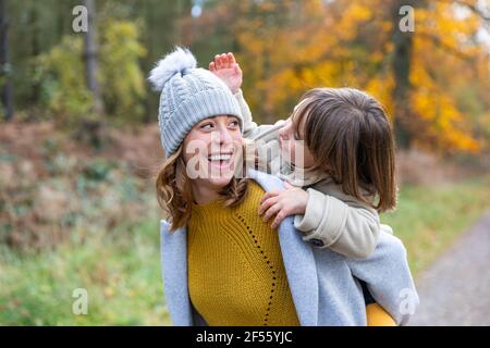 Cheerful mother piggybacking daughter while standing at forest Stock Photo