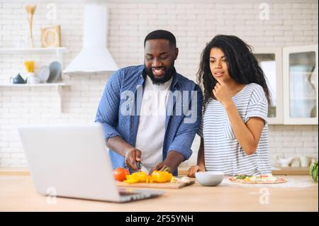 Happy african american spouses using laptop computer while cooking in modern kitchen together, searching recipe in internet, smiling black couple preparing healthy food for dinner at home, copy space Stock Photo