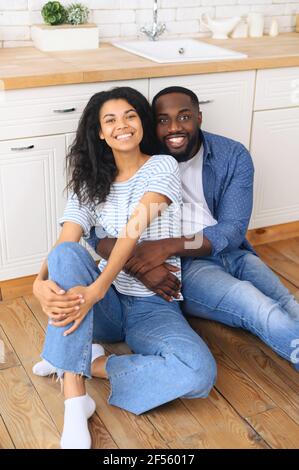 Tender loving young male african american husband sit on cozy wooden warm kitchen floor embrace hispanic wife hold her before him, happy lovers enjoying date, spending time together in new home Stock Photo