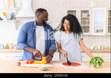Romantic couple is cooking on kitchen. Handsome man cuts vegetables and attractive young woman prepares the dough by spreading it with tomato paste, for dinner there will be an appetizing little pizza Stock Photo