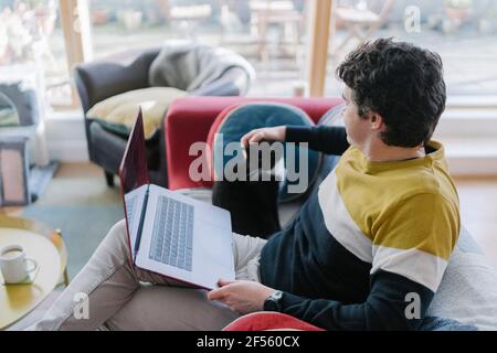 Mid adult man with laptop stroking cat while sitting on sofa at home Stock Photo
