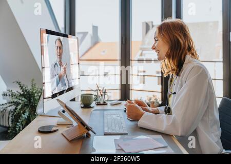 Female doctor talking with coworker over computer while sitting at office Stock Photo