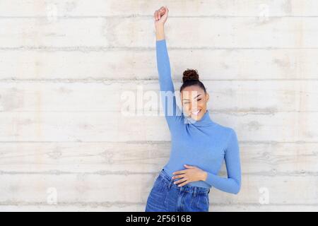 Young woman smiling while standing with hand raised against wall Stock Photo