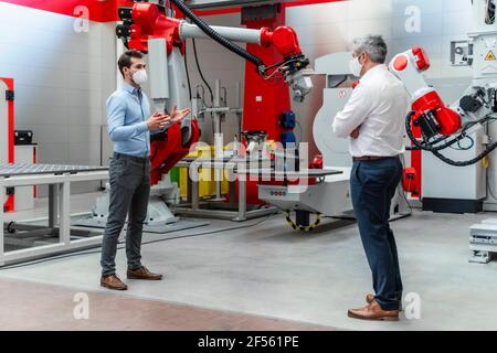 Young male entrepreneur with protective face mask discussing with colleague in factory Stock Photo