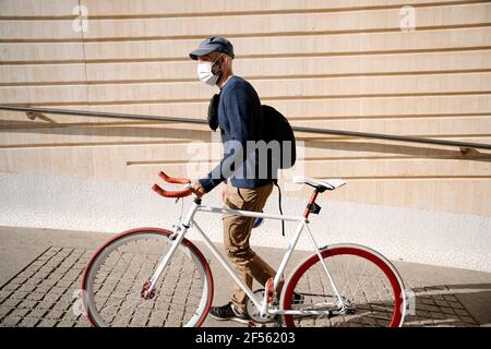 Man with bicycle looking away while walking by wall on footpath during COVID-19 Stock Photo