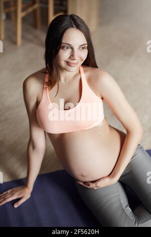 Vertical shot of cute happy european young pregnant woman in sports bra and leggings, leaning on hand while lying on roll pad, touching belly, feeling Stock Photo