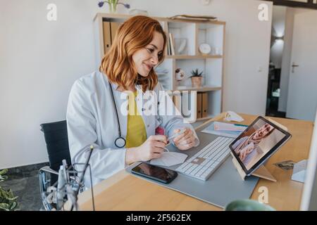 Doctor listening to patient on video call over digital tablet while sitting at office Stock Photo