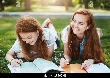 Stop fooling around and do homework. Two relaxed and carefree redhead sisters with freckles, lying on grass in park, writing notes for university Stock Photo