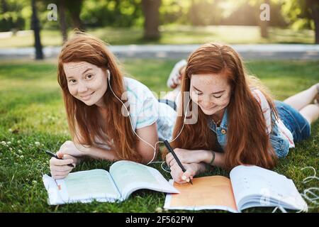 Homework are you kidding me. Two charming girls with red hair lying and chilling on grass during free time, doing homework, sister helping sibling Stock Photo