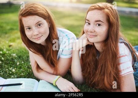 Close-up shot of two beautiful sisters with freckles and ginger hair, lying on grass, hanging out and making lessons, friend trying help girl with Stock Photo