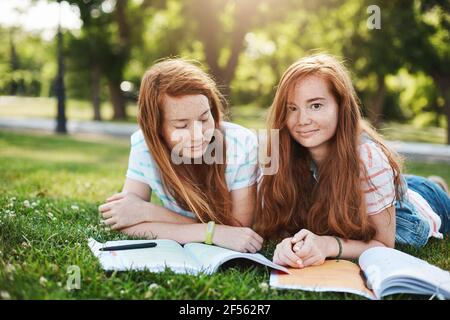 Friendship, family and lifestyle concept. Two attractive redhead girls relaxing on sunny day, lying on grass and doing homework together, smiling at Stock Photo