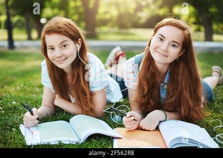 Sister trying to help sibling with lessons, being her personal tutor. Joyful and relaxed two girls with ginger hair and freckles, lying on grass while Stock Photo