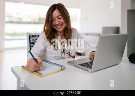 Smiling mature woman writing in diary while using laptop at home Stock Photo