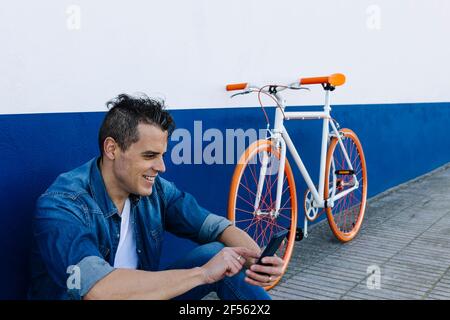 Smiling handsome man using mobile phone by bicycle against wall Stock Photo